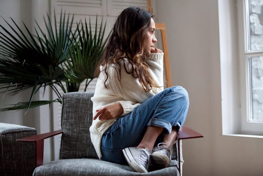 young woman sits on a chair and looks out a window while thinking about how anxiety and addiction are connected