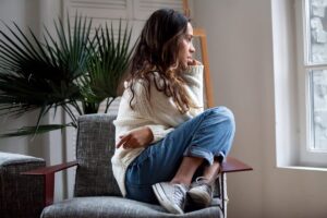 young woman sits on a chair and looks out a window while thinking about how anxiety and addiction are connected