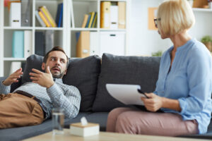 a man lays on a couch while a woman sits with him on the couch holding a piece of paper and discussing the importance of addressing underlying issues