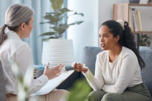 a woman sits on a couch looking at her therapist sitting across from her with notepad talking to the patient about the benefits of a residential treatment program