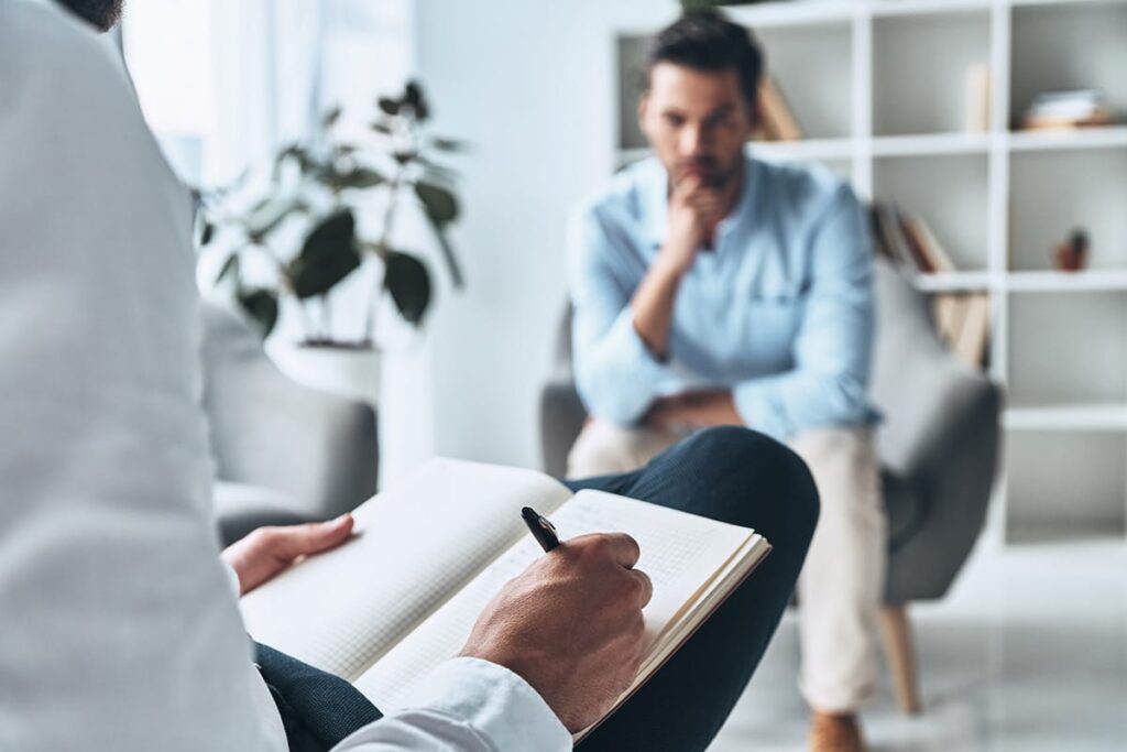 man sits on a chair and is thinking about what is dbt while his therapist takes notes while sitting across from him