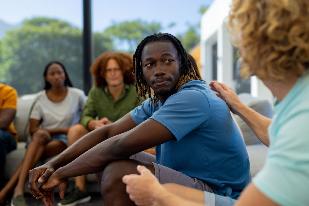 a group of people are sitting outside while a man puts his hand on the shoulder of another guy and answering his question about what is life skills training