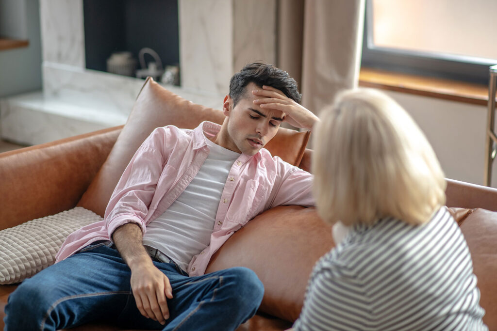 a distressed young adult male sits on a couch and asks the therapist sitting near him how does cognitive behavioral therapy work