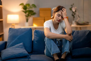 an upset woman sits on her couch with hand to her head wondering how long does it take to get addicted to alcohol